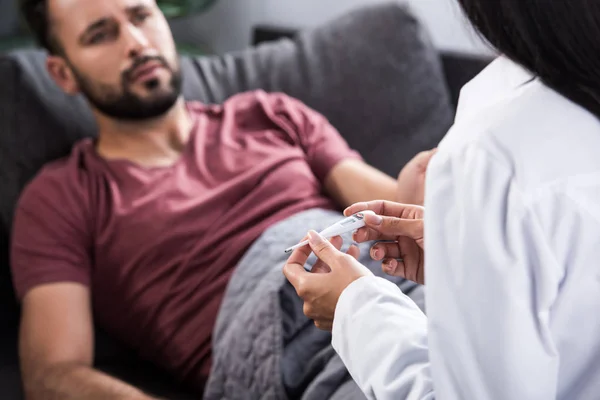 cropped shot of female doctor holding electric thermometer while her patient lying on couch on background