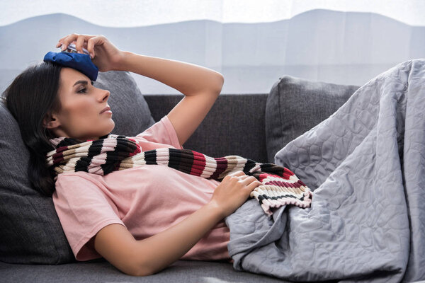sick young woman with high temperature holding ice pack on head while lying on couch