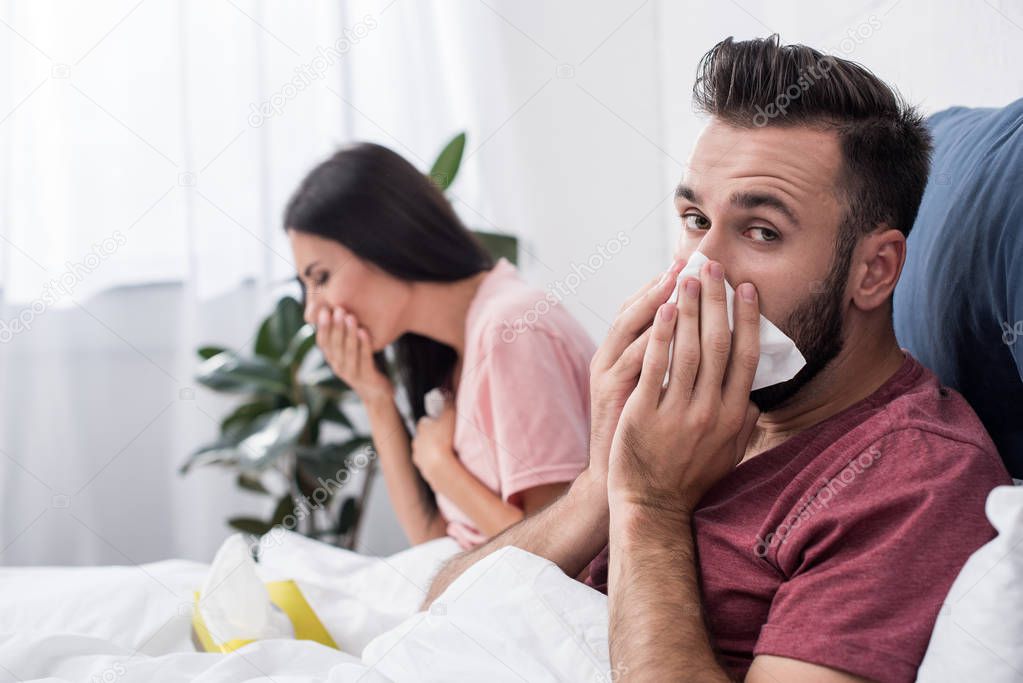 diseased young sick couple sneezing into paper napkins while sitting in bed