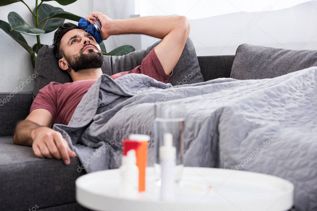 sick young man lying on couch and holding ice pack on forehead