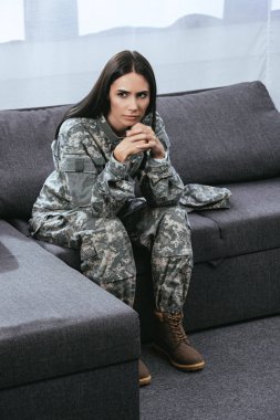 thoughtful female soldier in military uniform with ptsd sitting on couch and looking away clipart