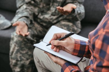 cropped shot psychiatrist writing notes during therapy session with female soldier clipart