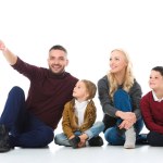 Rodina s rozkošný děti, které sedí na podlaze, otec dává něco izolované na bílém