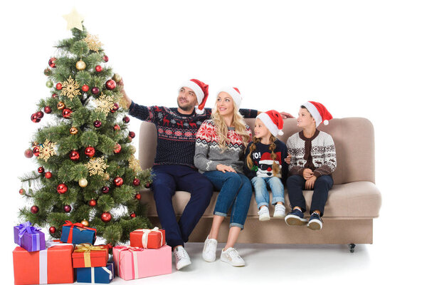 happy family in santa hats sitting on sofa near christmas tree with presents, isolated on white