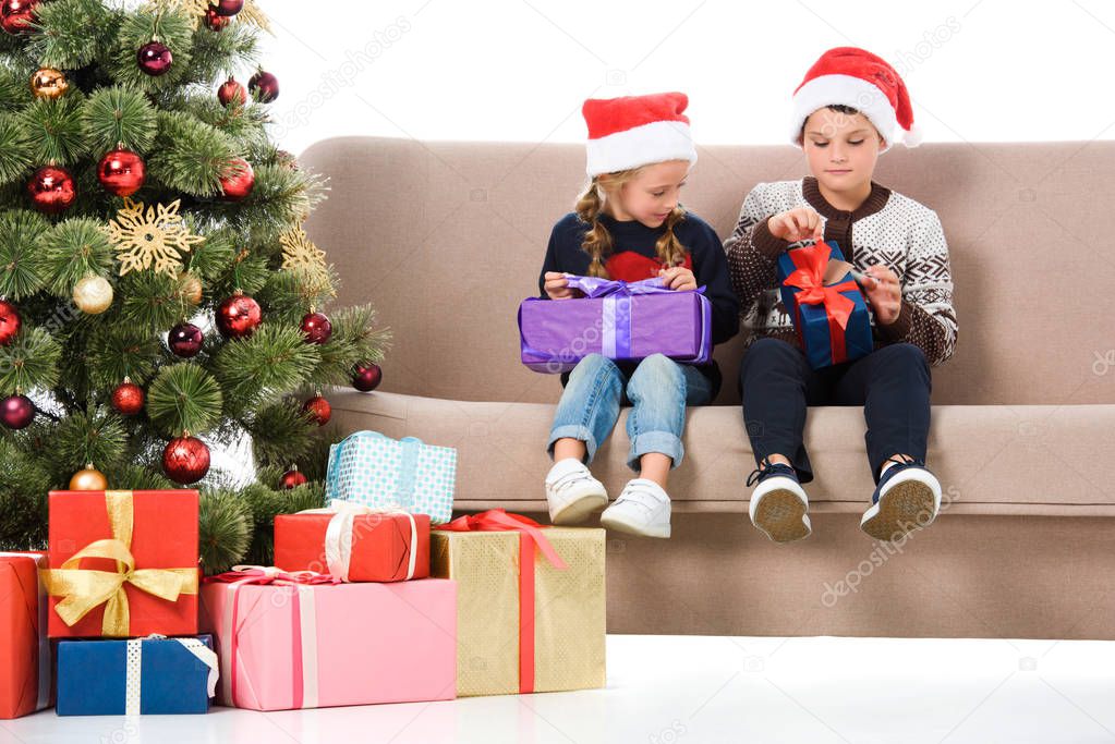 brother and sister in santa hat holding presents and sitting on sofa near christmas tree, isolated on white