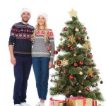 Beautiful happy couple in santa hats standing near christmas tree with gifts, isolated on white