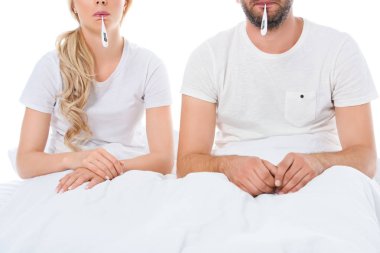 cropped view of sick couple with electronic thermometers sitting in bed, isolated on white clipart
