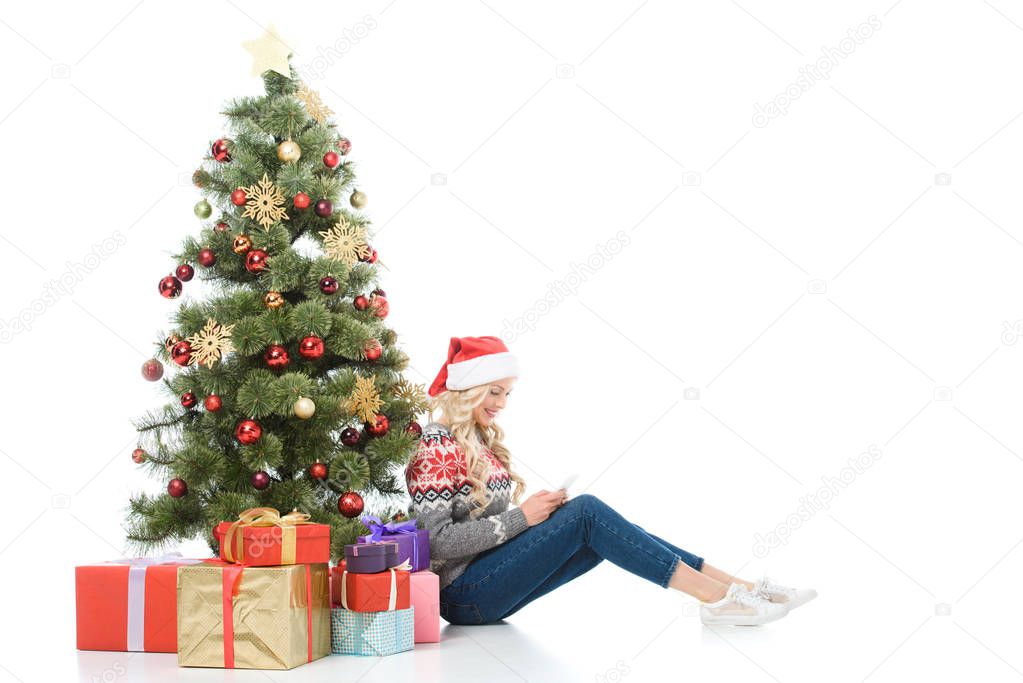 beautiful woman using smartphone and sitting near christmas tree with gifts, isolated on white