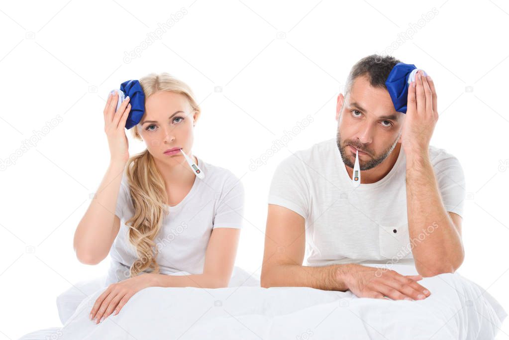 upset couple with electronic thermometers holding ice packs on heads and sitting in bed, isolated on white