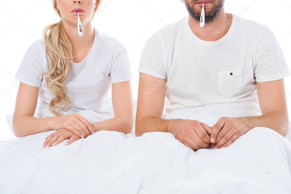 cropped view of sick couple with electronic thermometers sitting in bed, isolated on white