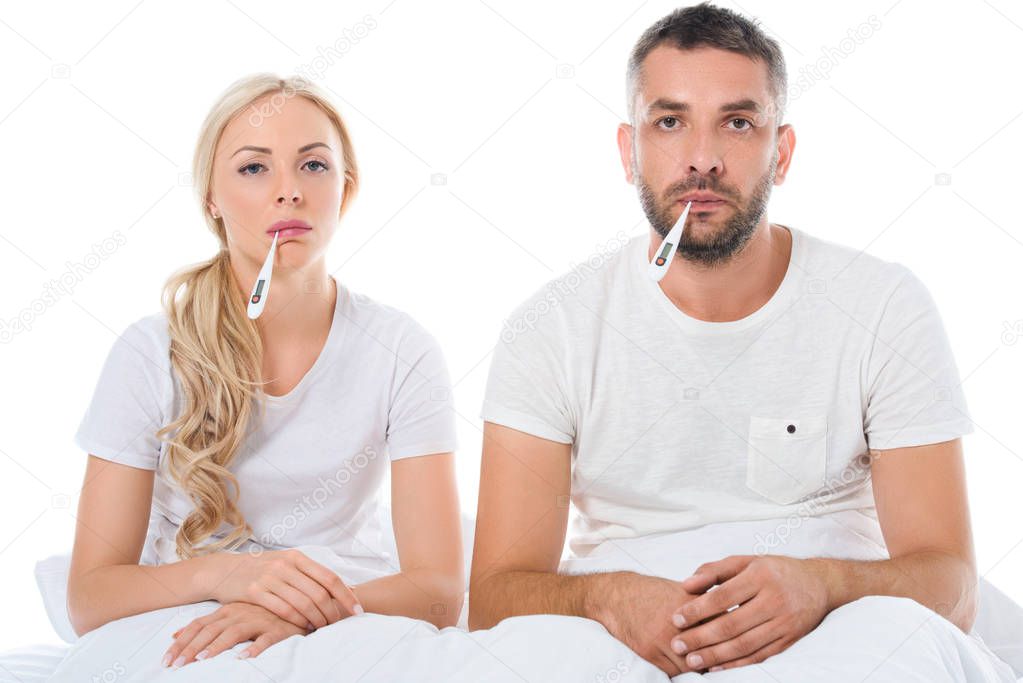 upset sick couple with electronic thermometers sitting in bed, isolated on white
