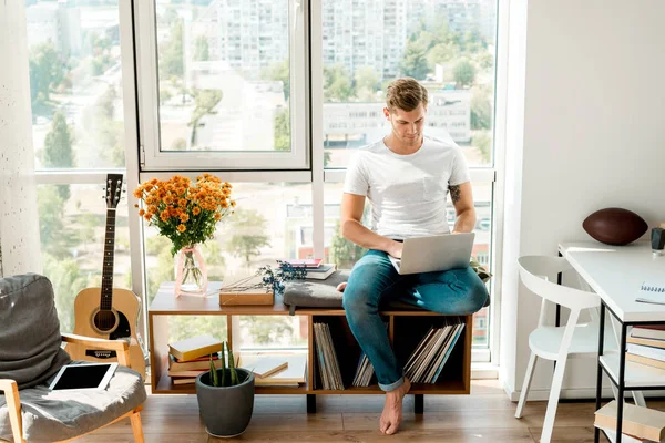 young man in casual clothing using laptop while sitting at window at home