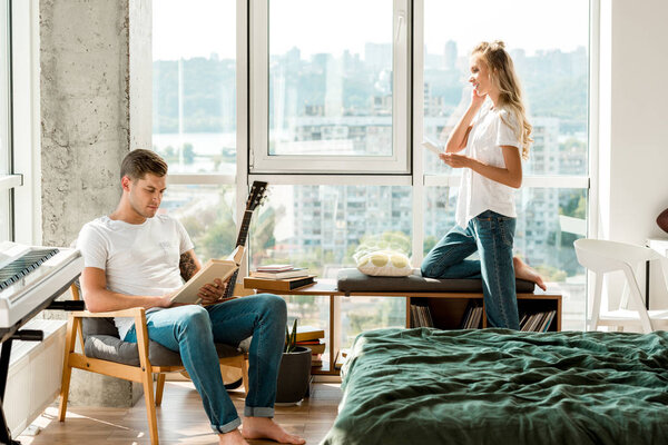 young woman in earphones listening music while boyfriend reading book in armchair at home