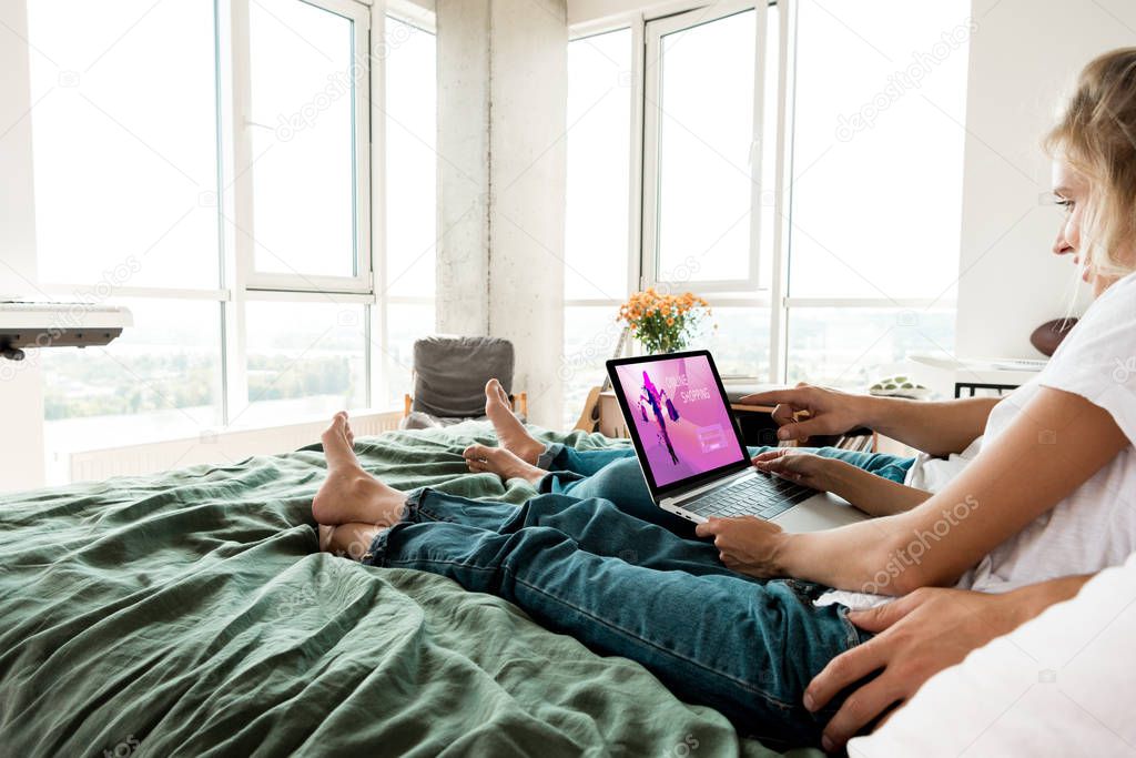 Partial view of couple with digital laptop with online shopping sign on screen resting on bed at home