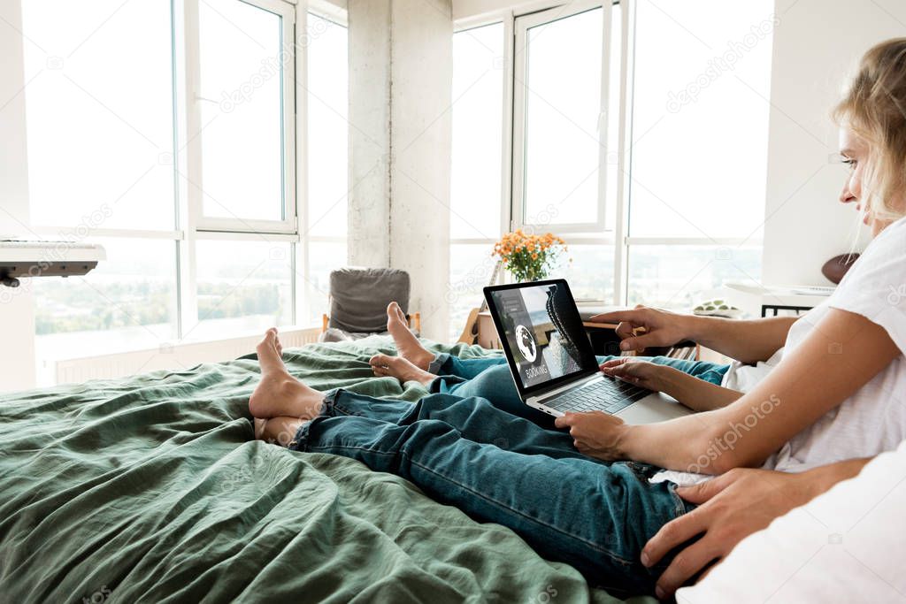 Partial view of couple with digital laptop with booking lettering on screen resting on bed at home