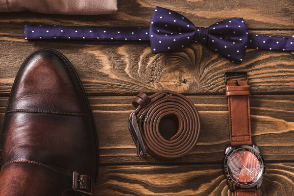 top view of leather male shoes, bow tie, belt and wristwatch arranged on wooden surface