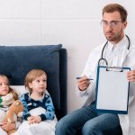 Handsome young pediatrician pointing at clipboard while sitting near kids lying in bed