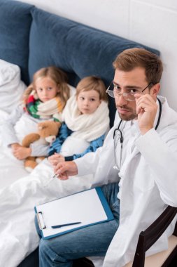 Pano ve yatakta yatan çocukların yanında otururken kameraya bakarak termometre ile yakışıklı çocuk doktoru yüksek açılı görünüş