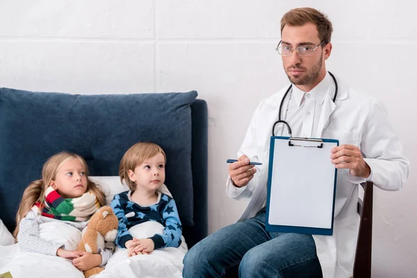 英俊的年轻儿科医生指着剪贴板 而坐在孩子们躺在床上 — 免费的图库照片