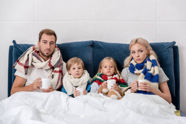 больная молодая семья с чашками горячего напитка сидит в постели и смотрит в камеру
