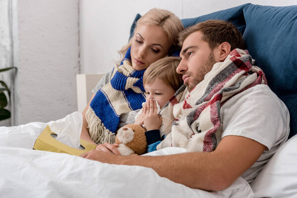 sick little kid blowing nose while lying in bed with parents