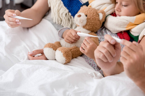 обрезанный снимок маленького больного ребенка, лежащего в постели с родителями и смотрящего на электрический термометр
