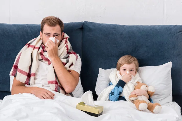 生病的父亲和儿子躺在床上 看着相机 用纸巾吹鼻子 — 图库照片