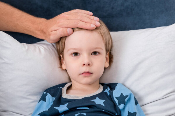 cropped shot of father checking sons temperature with hand while he lying in bed