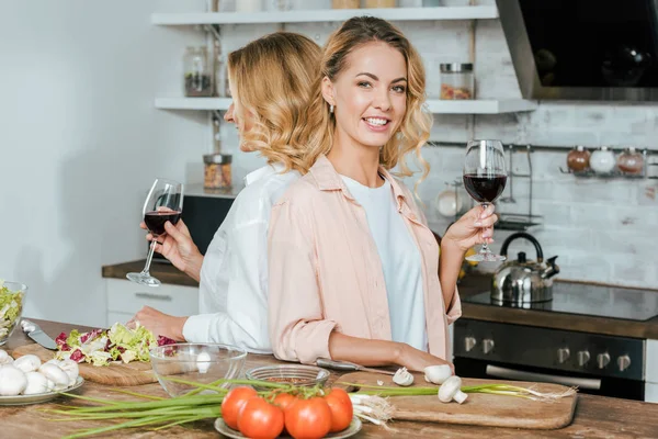 mature mother and smiling adult daughter with glasses of red wine standing back to back at kitchen