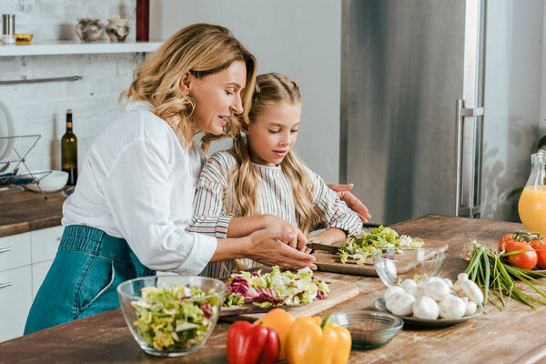 happy adult mother and little daughter cutting lettuce for salad together at home