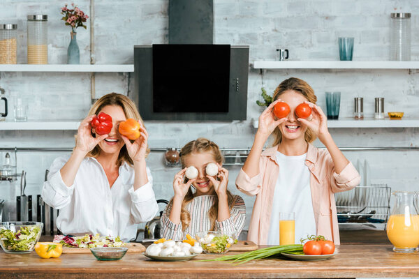 playful little child covering eyes with food with mother and grandmother and looking at camera while cooking