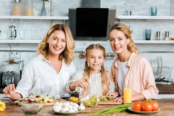 child cutting vegetables for salad with mother and grandmother at home and looking at camera