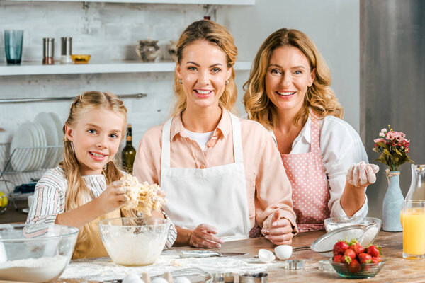 smiling child with mother and grandmother making dough together at home and looking at camera