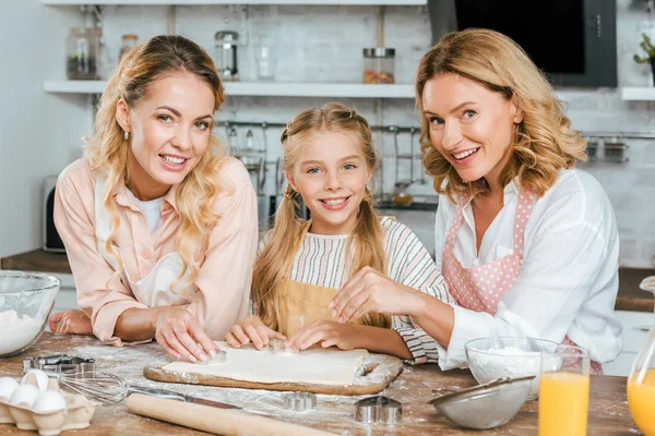 beautiful happy three generations of women making dough together at home and looking at camera