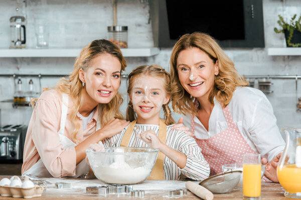 happy little child preparing dough with mother and grandmother at home and looking at camera