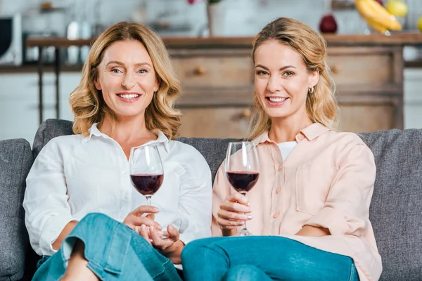 happy mature mother and adult daughter with glasses of red wine spending time together at home and looking at camera