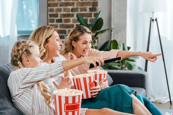 兴奋的孩子与母亲和祖母在家里看电影在沙发上的桶爆米花和指向屏幕 — 图库照片