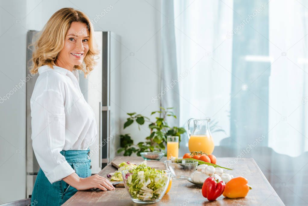 side view of happy adult woman looking at camera while making salad at kitchen