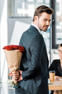 smiling handsome businessman hiding bouquet of roses behind back to surprise businesswoman in office clipart