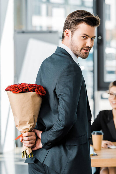 smiling handsome businessman hiding bouquet of roses behind back to surprise businesswoman in office