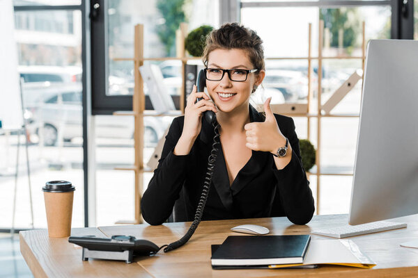 smiling attractive businesswoman talking by stationary telephone in office and showing thumb up