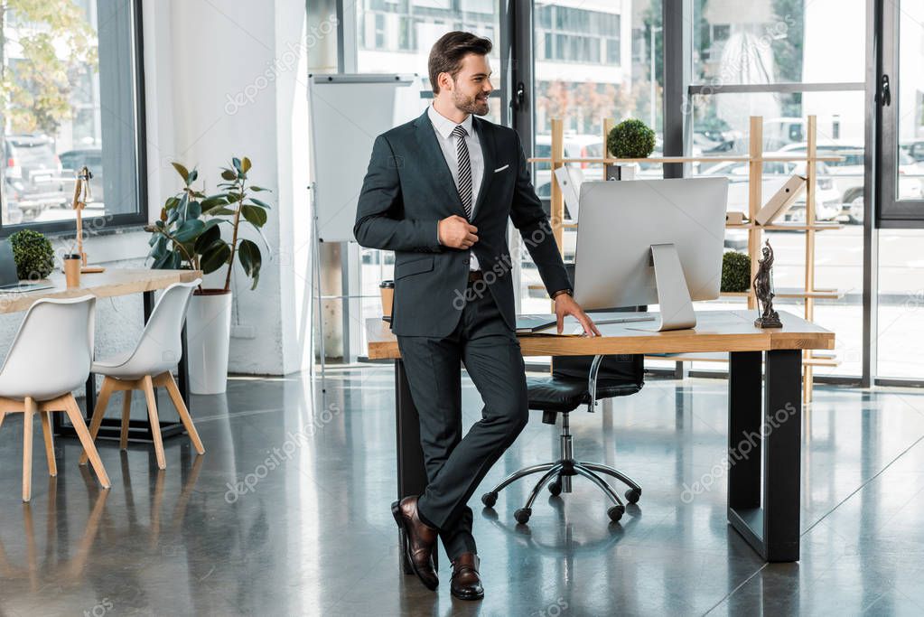 handsome businessman standing near table in office and looking away