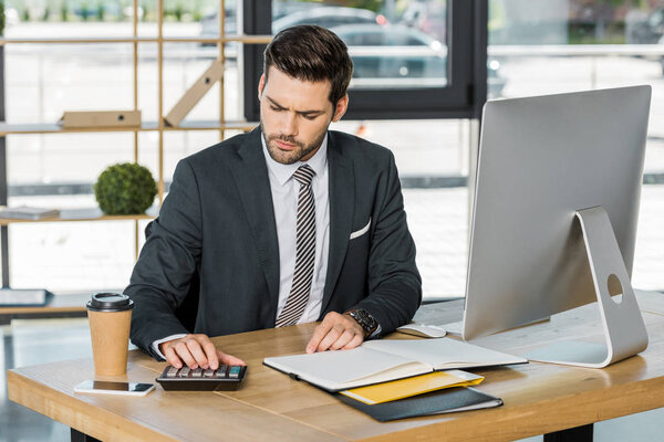 серьезный привлекательный бизнесмен с помощью калькулятора за столом в офисе
