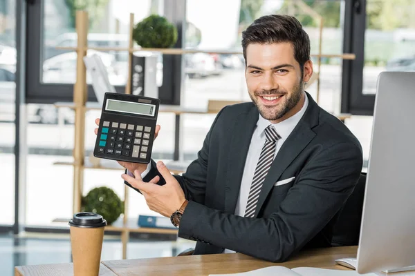 Lächelnder Gutaussehender Geschäftsmann Zeigt Taschenrechner Tisch Büro Und Blickt Die — kostenloses Stockfoto