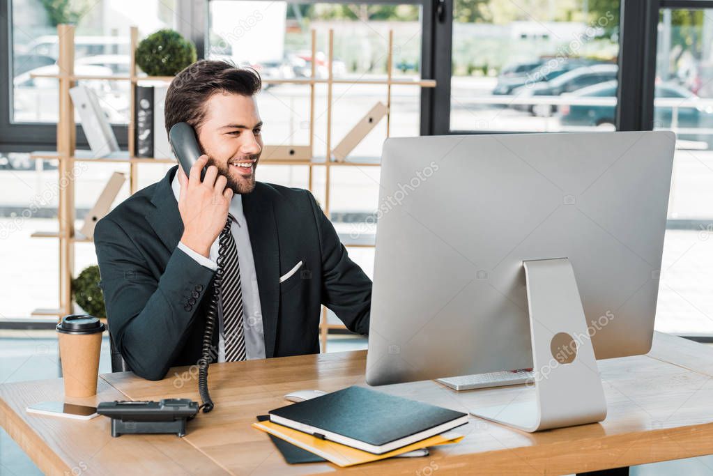 smiling handsome businessman talking by stationary telephone in office and looking at computer
