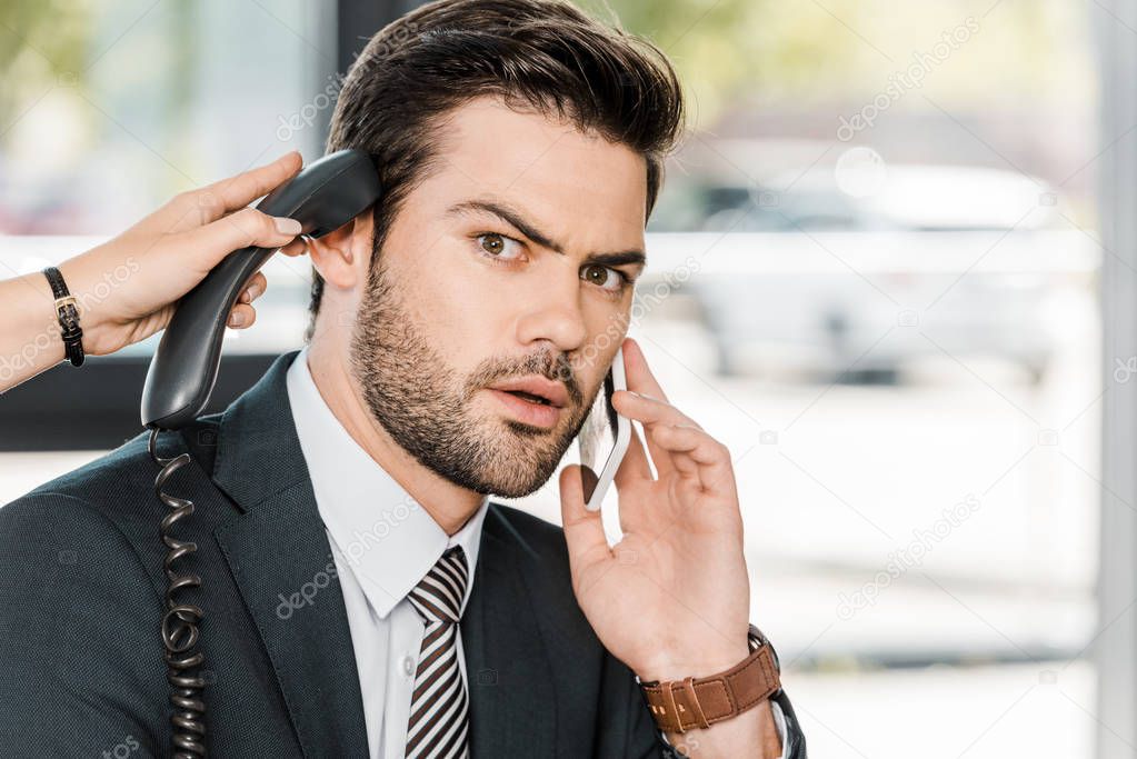 shocked businessman talking by smartphone and secretary giving him handset of stationary telephone