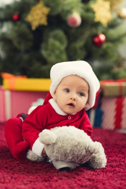 close-up portrait of cute little baby in santa suit lying on red carpet with teddy bear in front of christmas tree and gifts clipart