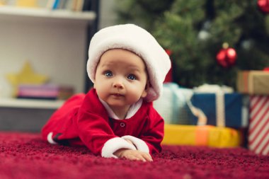 close-up portrait of beautiful little baby in santa suit lying on red carpet in front of christmas tree and gifts clipart