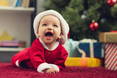 close-up portrait of laughing little baby in santa suit lying on red carpet in front of christmas tree and gifts clipart