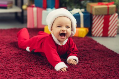 laughing little baby in santa suit lying on red carpet in front of christmas tree and gifts clipart